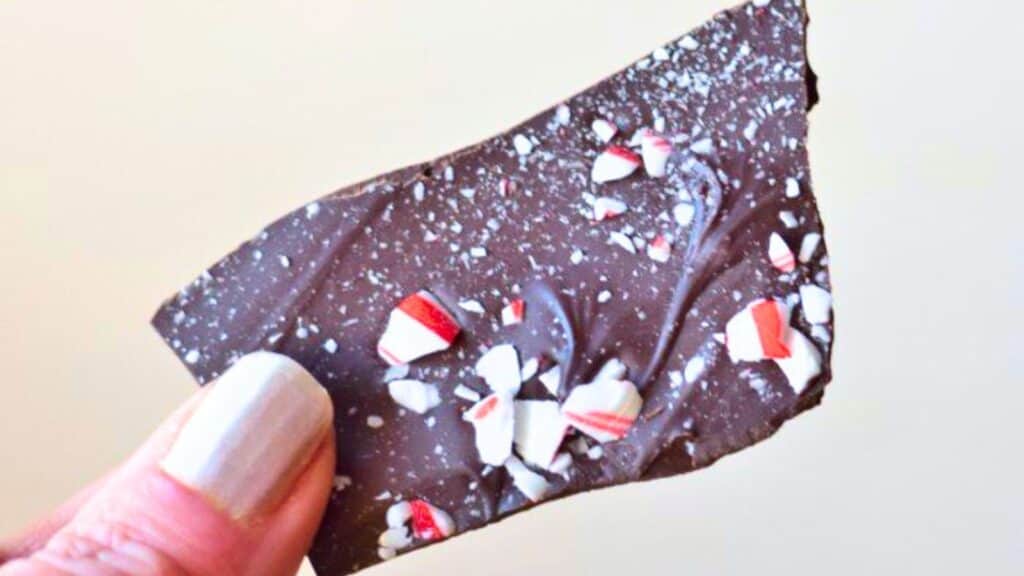 A hand holding a piece of dark chocolate bark topped with crushed candy canes.