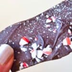 A hand holding a piece of dark chocolate bark topped with crushed candy canes.