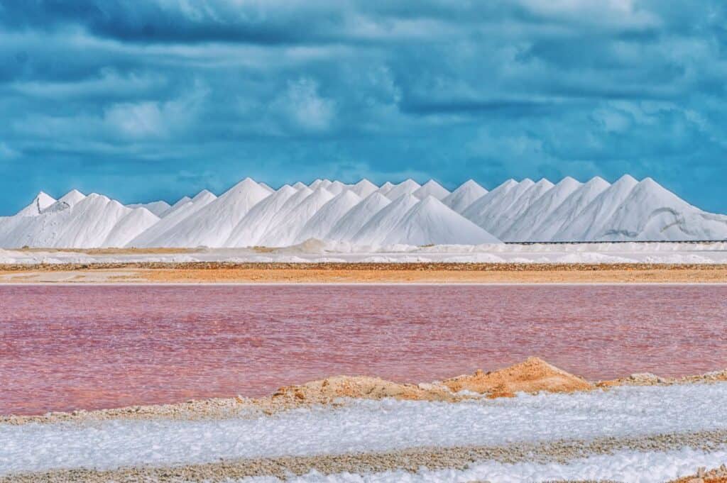 White salt mounds under a blue sky with pink-hued salt ponds and a foreground of sandy and salt terrain.