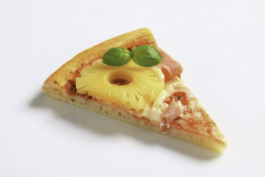 A slice of pizza topped with a ring of pineapple, pieces of ham, melted cheese, and garnished with two basil leaves on a plain white background.