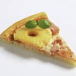 A slice of pizza topped with a ring of pineapple, pieces of ham, melted cheese, and garnished with two basil leaves on a plain white background.