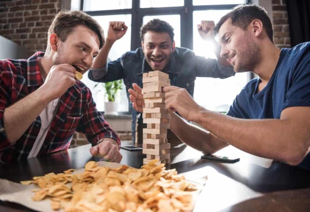 Three men are playing a game of Jenga at a table with snacks. One man places a block while another bites his nails, and the third man watches excitedly with his arms raised.