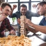 Three men are playing a game of Jenga at a table with snacks. One man places a block while another bites his nails, and the third man watches excitedly with his arms raised.