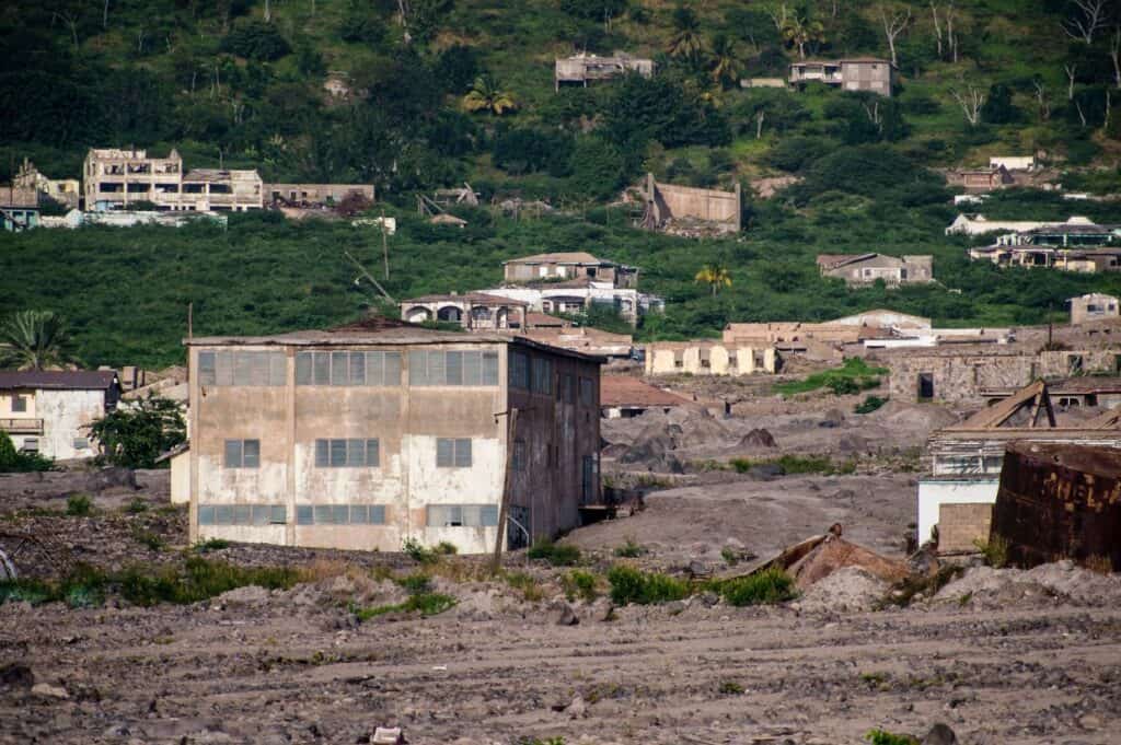 A weathered building stands among deserted, partially ruined structures on a green hillside, surrounded by barren, rocky ground—an evocative scene emblematic of tourism in Montserrat.