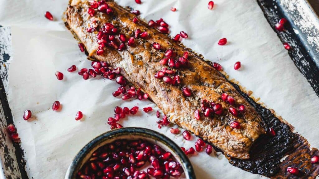 Grilled fish fillet garnished with pomegranate seeds on parchment paper, with a small bowl of pomegranate seeds beside it.