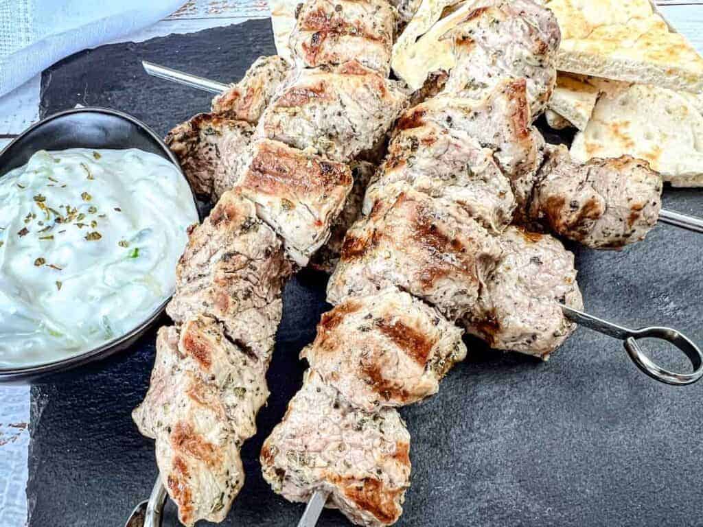 Three pieces of Pork Souvlaki with a bowl of white sauce and pieces of flatbread on a black slate serving board.