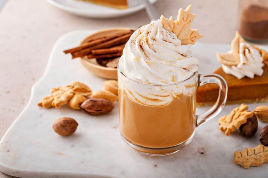 A glass mug filled with a pumpkin spice drink topped with whipped cream and a decorative leaf-shaped cookie. Surrounding the mug are autumn-themed cookies, cinnamon sticks, and nuts on a marble surface.