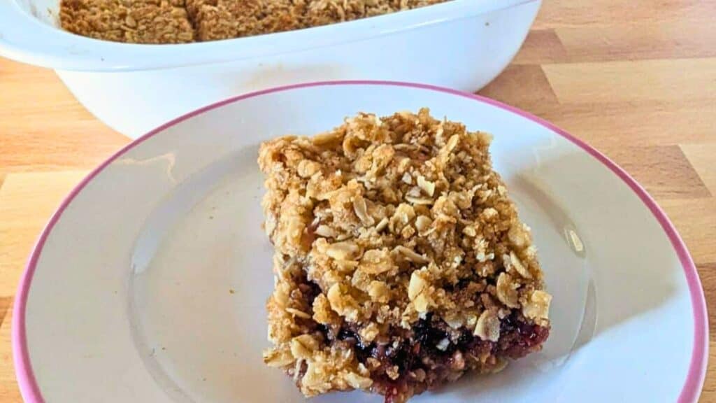 A square of a berry jam bar sits on a white plate with a pink rim. A white baking dish with the remaining crumble is in the background on a wooden surface.