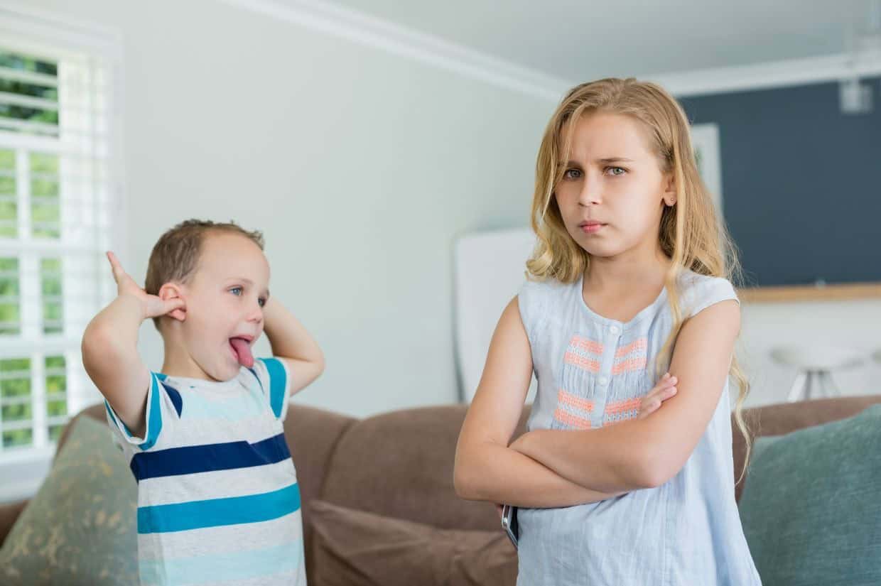 A young girl stands with her arms crossed and a displeased expression, while a boy behind her sticks out his tongue and places his hands on his ears playfully.