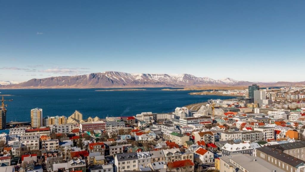 Panoramic view of Reykjavik, Iceland, with colorful buildings in the foreground, a clear blue sea, and snow-capped mountains in the background under a clear sky—a perfect highlight for your 4-day Iceland itinerary.