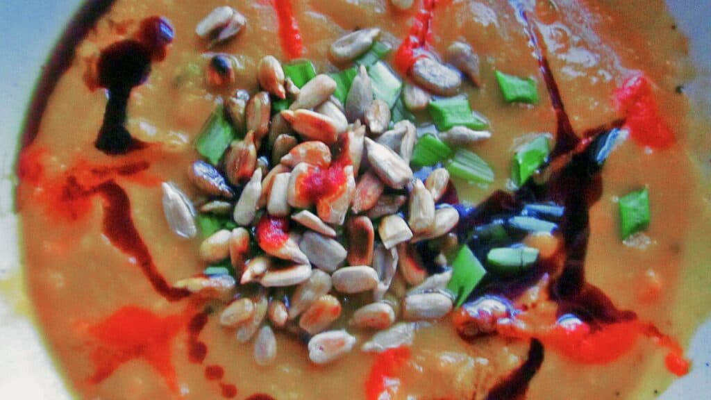 Close-up of a creamy soup garnished with sunflower seeds, chopped green onions, and drizzled with red and dark sauces.