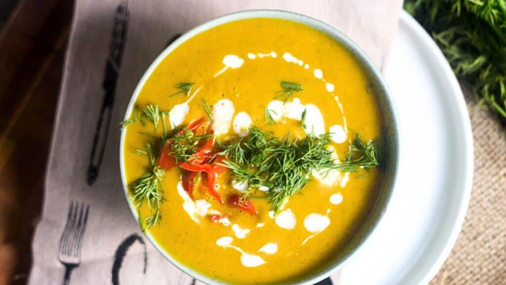 A bowl of creamy orange soup garnished with fresh dill, red peppers, and a drizzle of white sauce, placed on a folded napkin with cutlery print, next to a green herb on the side.