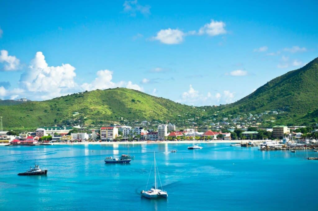 A coastal town with buildings at the base of green hills, several boats anchored in clear blue water under a partly cloudy sky, reminiscent of the exclusivity of Anguilla.