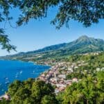 A coastal town with numerous buildings and boats in a harbor, surrounded by lush greenery reminiscent of Martinique's lush landscapes, and overlooked by a tall, forested mountain under a clear blue sky.