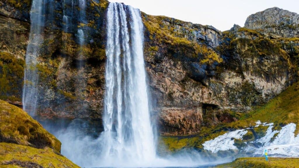 A large waterfall cascades down a rocky cliff with moss-covered surfaces, surrounded by patches of greenery and a small area of snow. A person in a blue jacket stands nearby for scale, making it an ideal highlight in any 4-day Iceland itinerary.