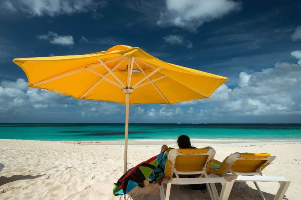 A person sits under a large yellow umbrella on a sandy beach with turquoise water and partly cloudy skies, embodying the exclusivity of Anguilla. Nearby, two white lounge chairs and colorful towels complete this serene paradise.