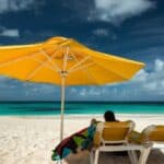 A person sits under a large yellow umbrella on a sandy beach with turquoise water and partly cloudy skies, embodying the exclusivity of Anguilla. Nearby, two white lounge chairs and colorful towels complete this serene paradise.