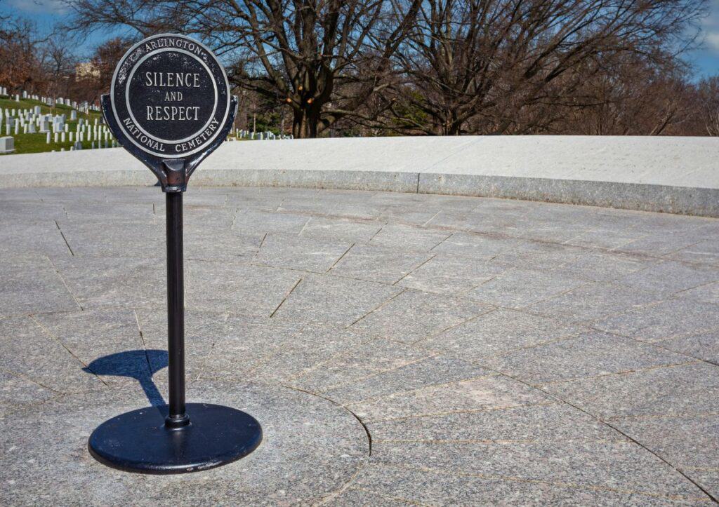 A sign at Arlington National Cemetery reads "Silence and Respect" on a stone-paved area, with grave markers and bare trees visible in the background, serving as a poignant reminder of Arlington National Cemetery facts.