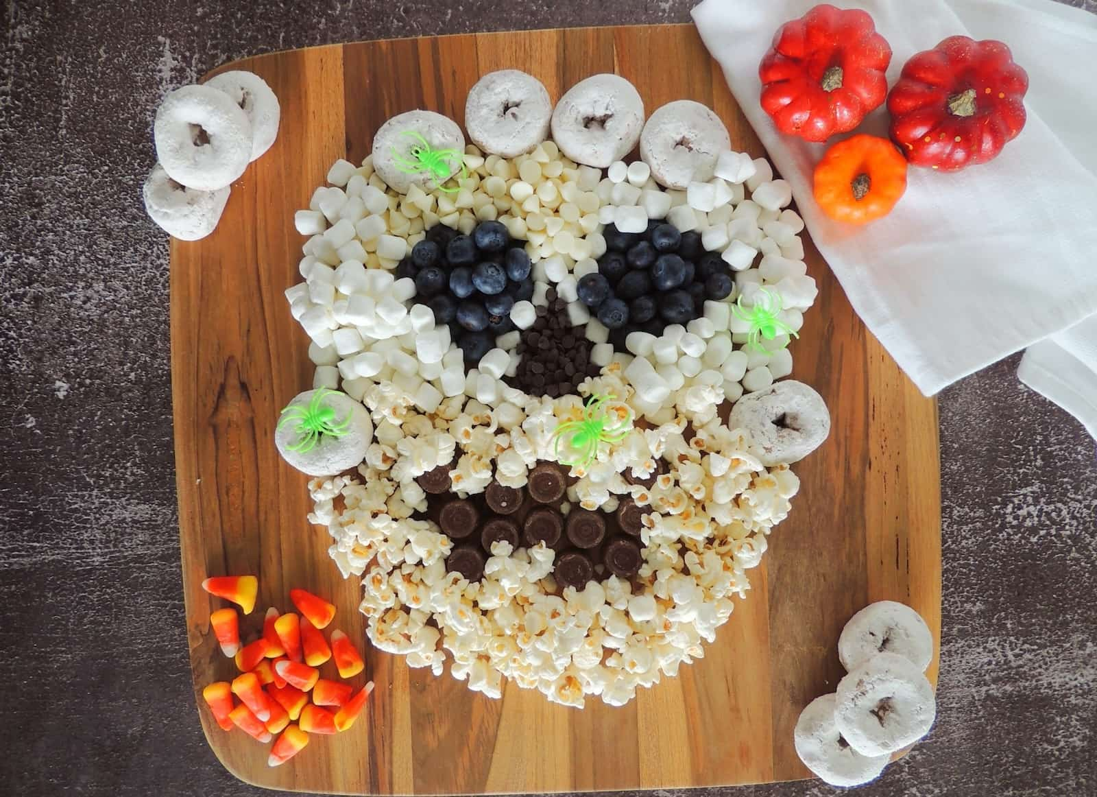 A Halloween-themed skull made of popcorn, marshmallows, blueberries, candy, and mini donuts on a wooden board, with candy corn and mini pumpkins as decorations.