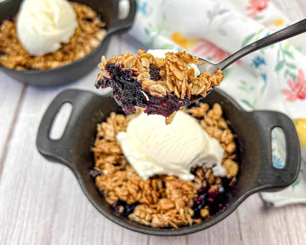 Close-up of a spoonful of blueberry crisp topped with ice cream, lifted above a black dish containing the same dessert with a scoop of ice cream in the background—a perfect addition to your backyard BBQ menu.