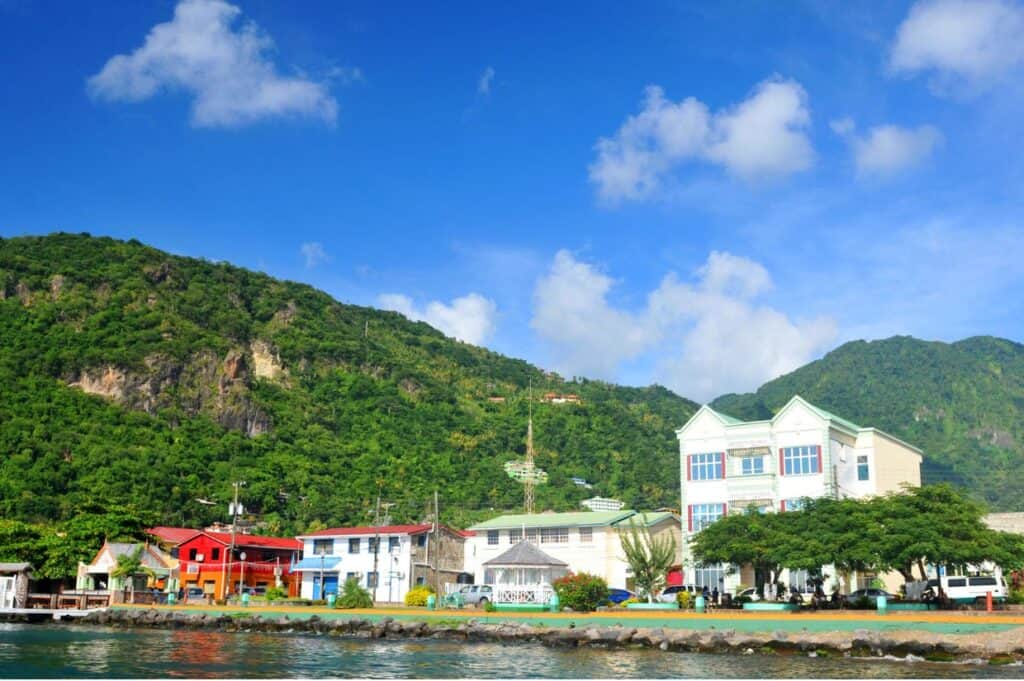 A coastal town with colorful buildings, lush green hills in the background, and blue skies above.