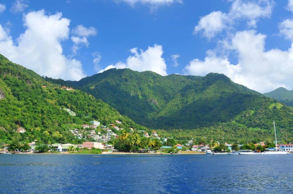 View of a coastal town with lush green hills in the background and boats docked near the shore under a clear blue sky.