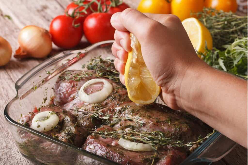 A hand squeezes a lemon over seasoned raw meat in a glass baking dish, surrounded by tomatoes, onions, and herbs.