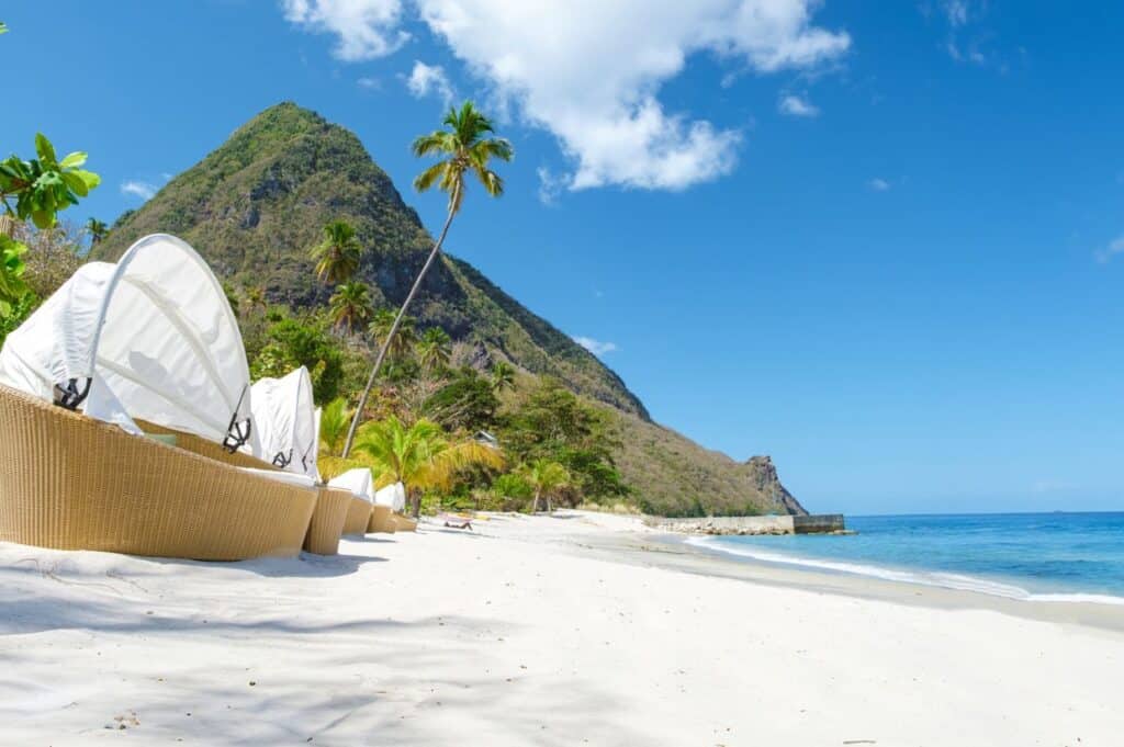 A tropical beach scene with white sand, wicker lounge chairs, palm trees, and a mountainous background under a clear blue sky.