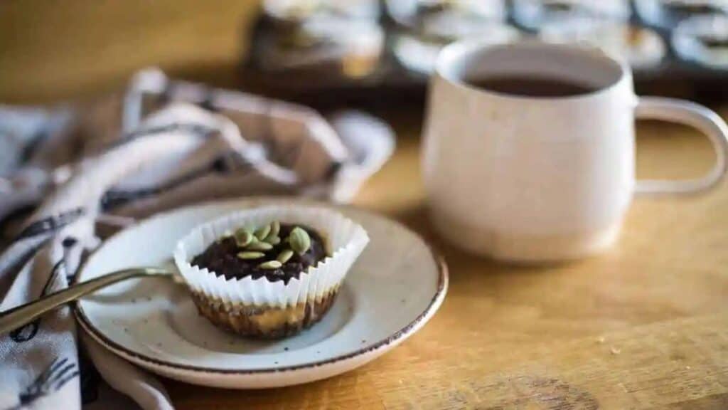 A white mug filled with coffee sits on a wooden table next to a small dessert in a white paper wrapper on a plate with a fork. A brown and white cloth is partially visible beside the plate.