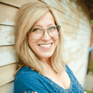 A woman with blonde hair and glasses smiles while leaning against a wooden wall.