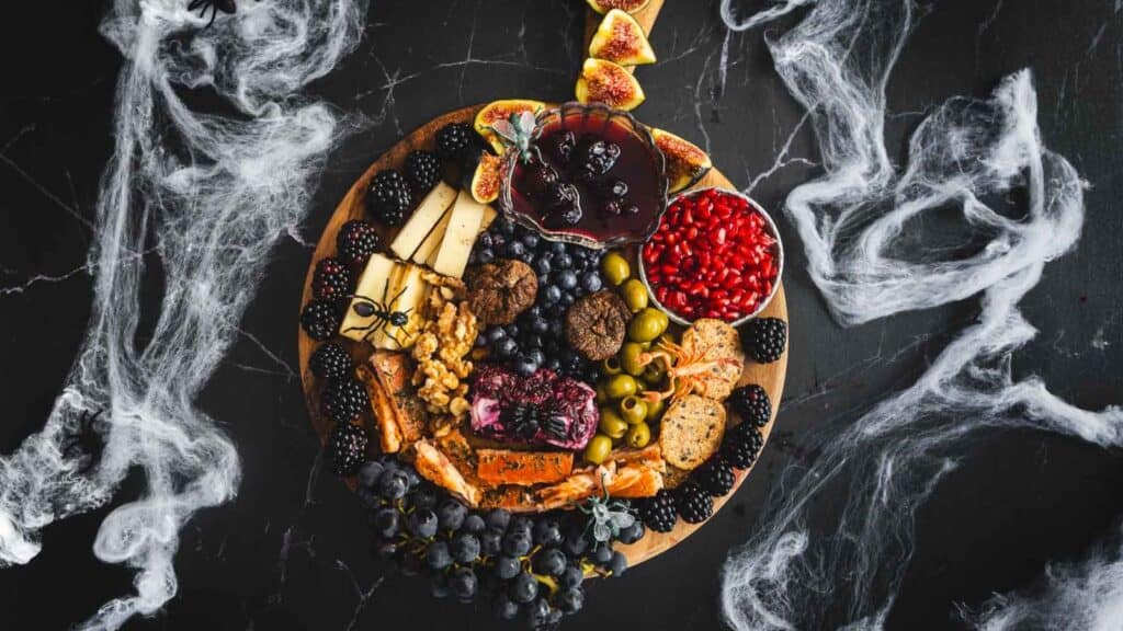 A round wooden board with an assortment of cheeses, crackers, fruits, olives, nuts, and a bowl of red dip. The board is surrounded by white smoke-like decorations on a dark surface.