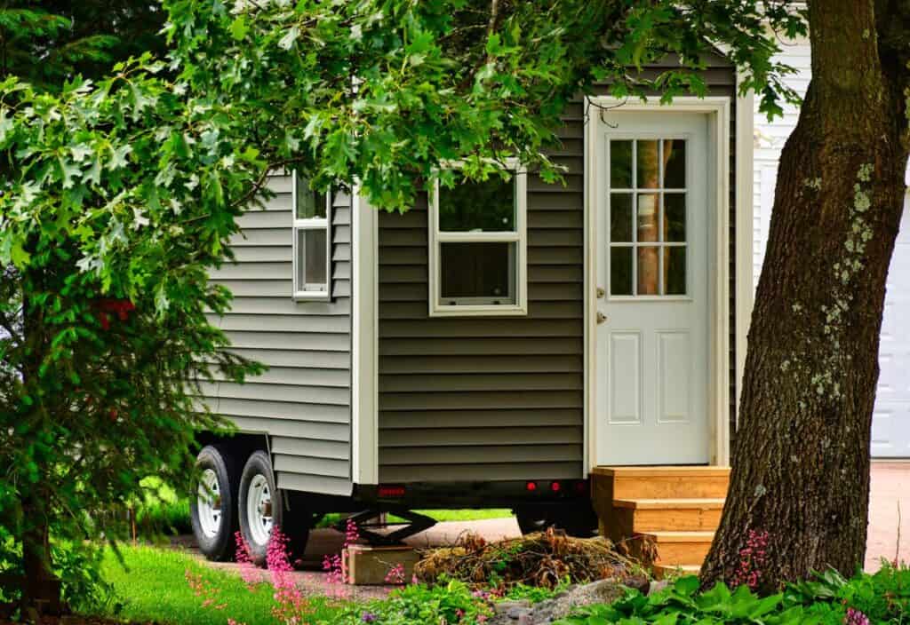 A tiny house on wheels with gray siding and a white door is parked in a green yard, partially hidden by trees.