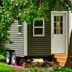 A tiny house on wheels with gray siding and a white door is parked in a green yard, partially hidden by trees.