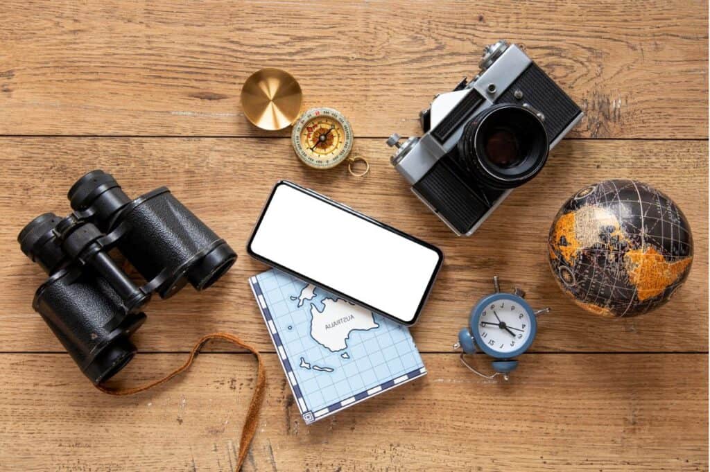 A flat lay of travel items: binoculars, a map, a compass, a vintage camera, a smartphone, a small globe, and an alarm clock on a wooden surface.