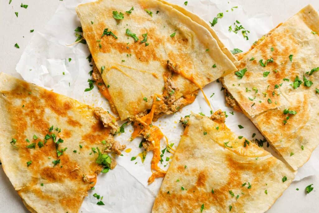 Four quesadilla slices on parchment paper, filled with melted cheese and tuna, garnished with chopped parsley.