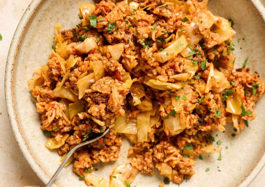 A bowl of cooked rice mixed with ground meat, cabbage, and garnished with chopped parsley. A fork is resting on the side of the bowl.