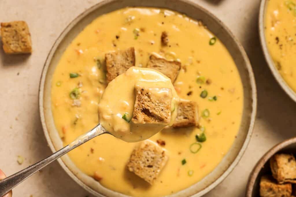 A bowl of creamy soup garnished with croutons and chopped green onions, with a spoonful of soup and croutons being lifted from the bowl.