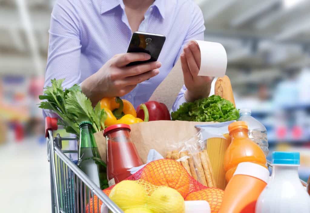 Person holding a receipt and a smartphone in a grocery store, with a shopping cart full of various groceries including vegetables, fruits, beverages, and packaged goods.