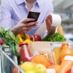 Person holding a receipt and a smartphone in a grocery store, with a shopping cart full of various groceries including vegetables, fruits, beverages, and packaged goods.