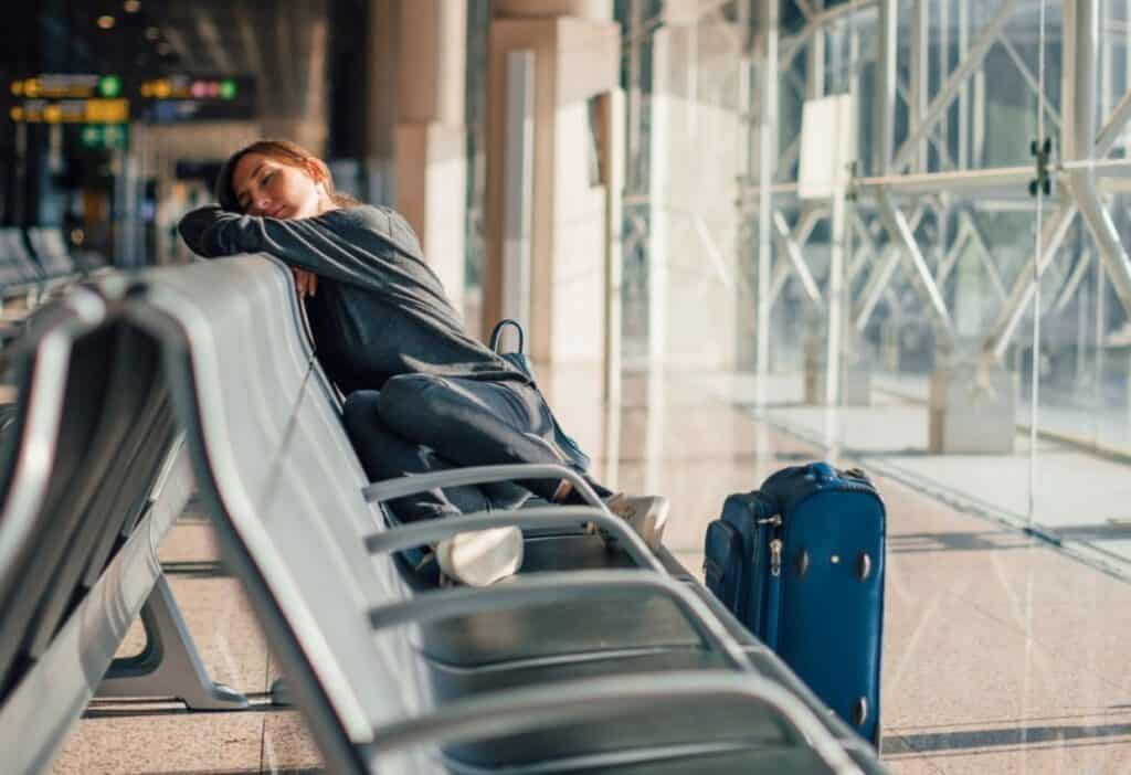 A person rests on a row of chairs in an airport terminal, with their head on their arms and a blue suitcase beside them.