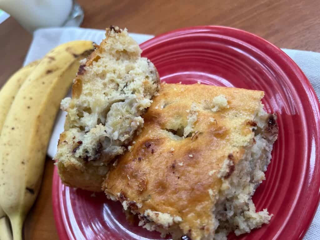 Two pieces of banana bread on a red plate, with a bunch of bananas and a glass of milk in the background.