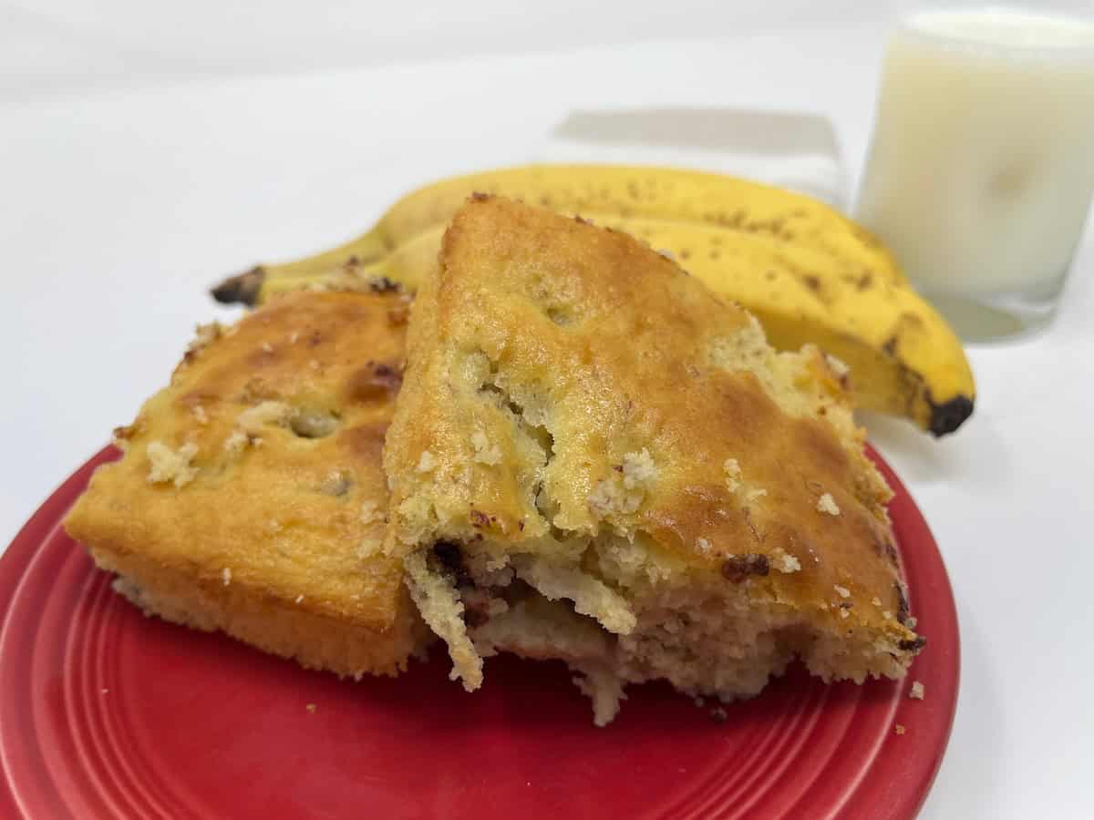 Two slices of banana bread on a red plate, with two ripe bananas and a glass of milk in the background.