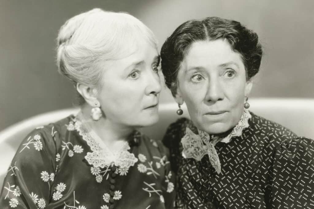 Two elderly women with concerned expressions, dressed in vintage clothing with lace collars, sit closely together. One leans in as if listening intently to the other.