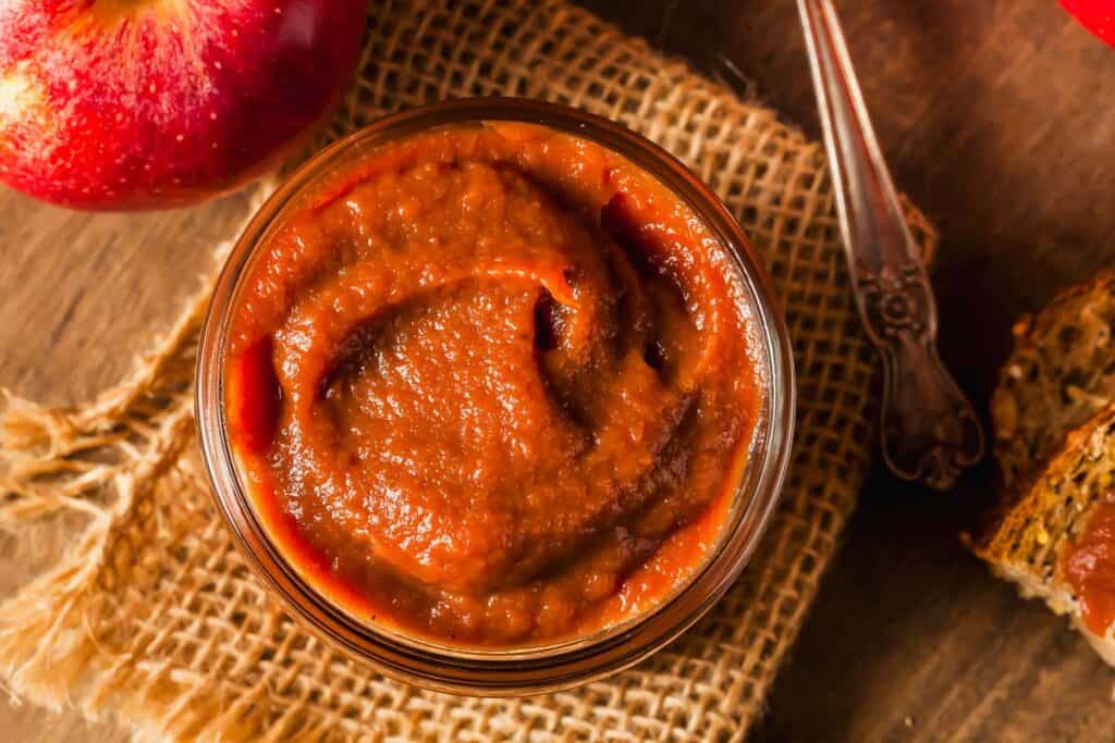 A jar of apple butter sits on a piece of burlap next to a spoon, an apple, and a slice of bread coated with apple butter.