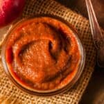 A jar of apple butter sits on a piece of burlap next to a spoon, an apple, and a slice of bread coated with apple butter.