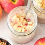 Two jars of apple oatmeal topped with chopped apples, surrounded by whole apples and a bowl of mixed nuts and dried fruits.