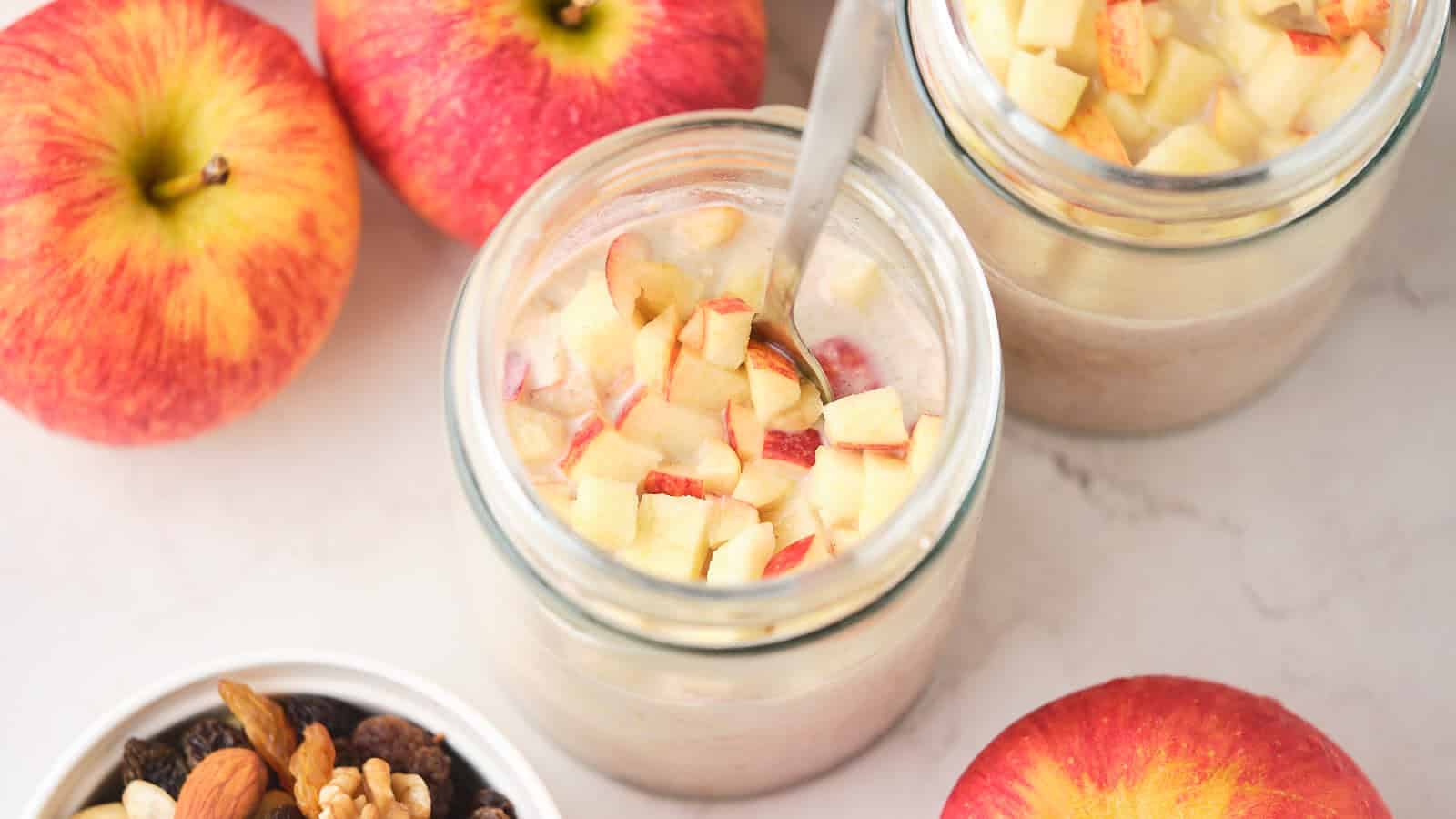 Two jars of apple oatmeal topped with chopped apples, surrounded by whole apples and a bowl of mixed nuts and dried fruits.