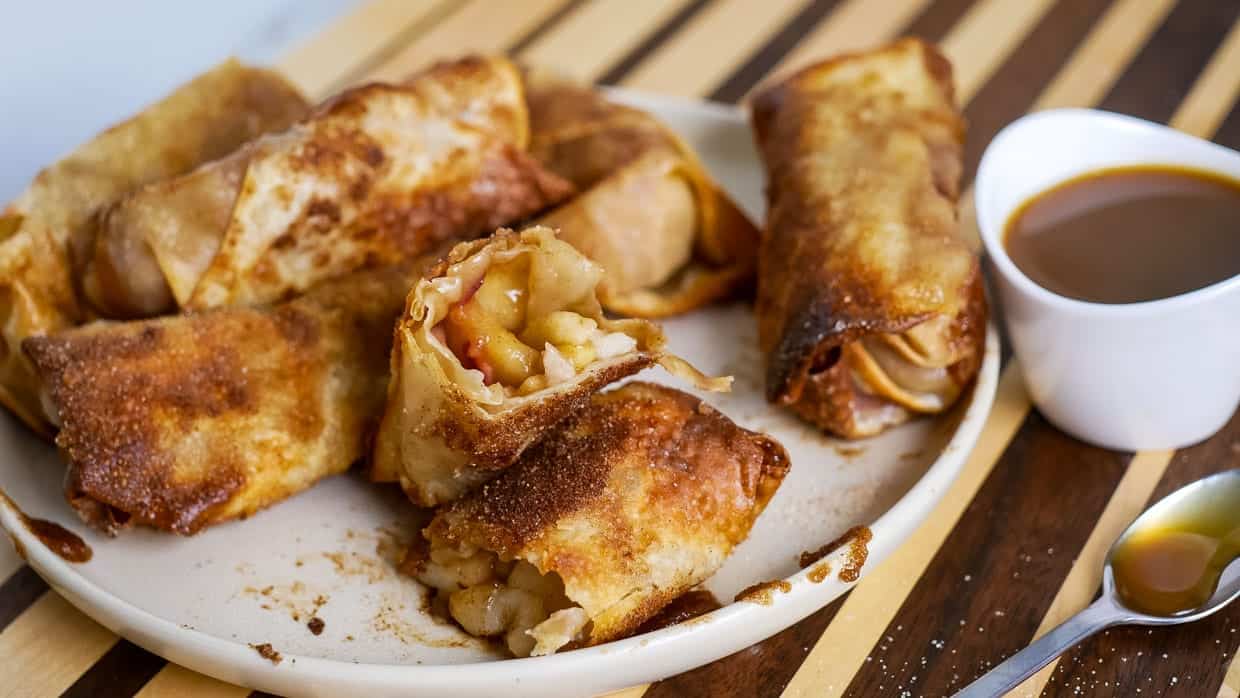 Plate of fried apple spring rolls served with a small bowl of caramel dipping sauce and a spoon on the side.