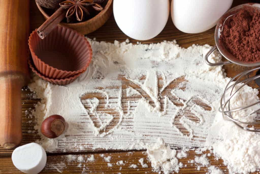 The word "Bake" written in flour surrounded by baking ingredients like eggs, cocoa powder, cinnamon sticks, star anise, a rolling pin, and baking cups on a wooden surface.