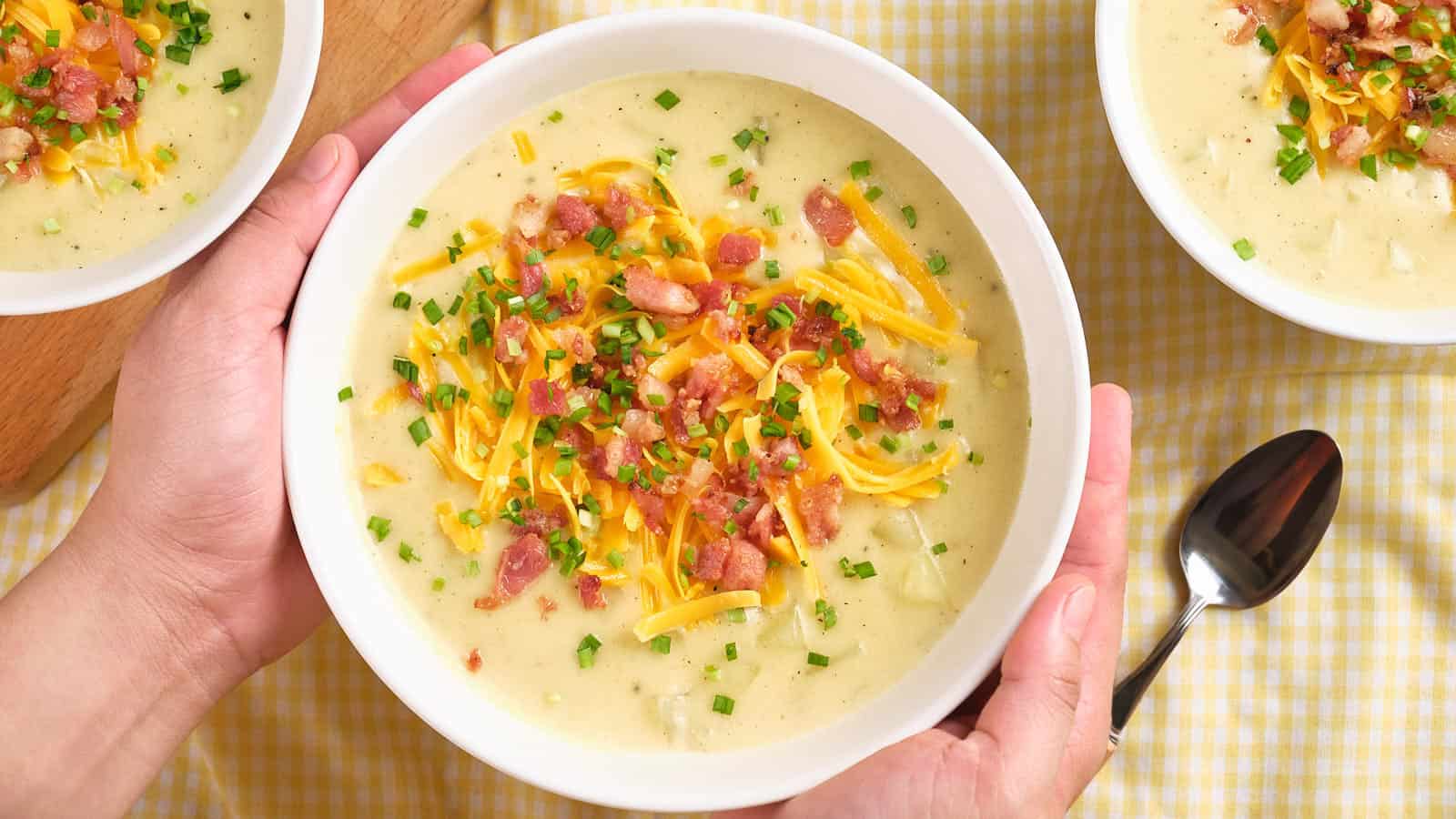 A person holding a bowl of baked potato soup garnished, with similar bowls and a spoon on a checkered tablecloth.